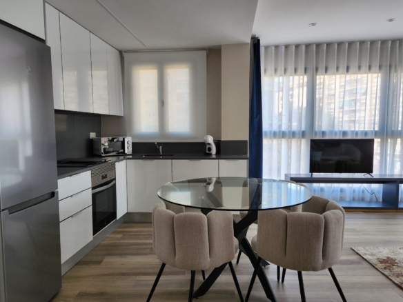 Cocina abierta moderna en tonos blancos y negros con mesa redonda de cristal y sillas tapizadas, junto a salón iluminado por ventanales amplios