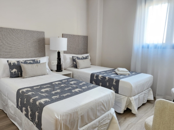 Dormitorio doble con dos camas individuales vestidas en tonos blancos y grises, cabeceros tapizados y ventana con cortinas claras