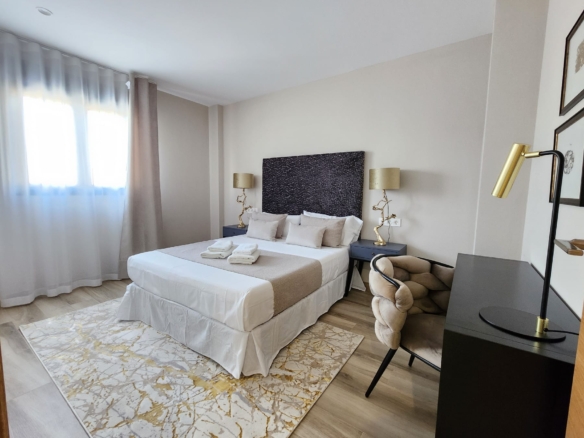 Dormitorio principal moderno con cama doble, cabecero texturizado oscuro, mesas con lámparas doradas y ventana con cortinas claras