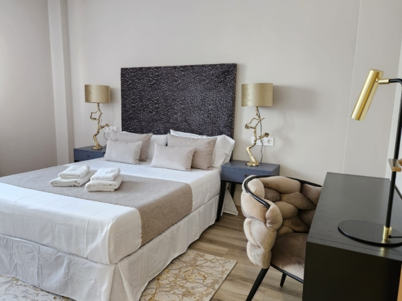 Dormitorio moderno con cama doble vestida en tonos neutros, cabecero oscuro texturizado, lámparas doradas y escritorio con silla acolchada